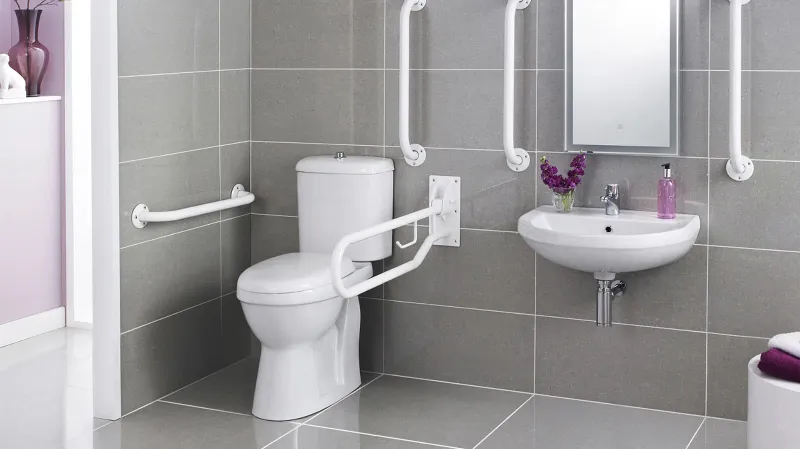 A wheelchair or handicap accessible bathroom with a toilet and sink featuring multiple white grab rails on grey tiled walls.
